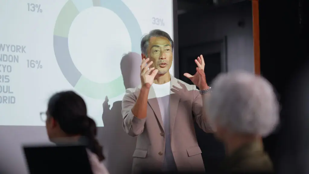 An Asian senior adult professional delivers a strategic presentation, utilizing data visuals and expressive gestures to convey key insights and financial metrics essential for effective business planning and decision-making